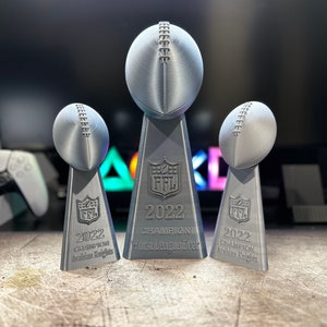 Op de afbeelding: Drie zilveren voetbaltrofee&euml;n met de tekst "FFL 2022 Champion" op elk. De middelste trofee heeft de tekst "Worst League Ever". De trofee&euml;n staan op een houten oppervlak.
