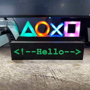 Op de afbeelding: Een zwart houten bord met de tekst "<!---Hello--->" in groene letters. Het bord staat voor vier gekleurde lampen in de vorm van een driehoek, cirkel, X en vierkant.