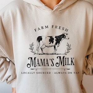 Op de afbeelding: Een cr&egrave;mekleurige hoodie met een zwart-witte koe-illustratie en de tekst "Farm Fresh Mama's Milk Locally Sourced - Always on Tap".