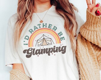 Peut inclure: Un t-shirt blanc avec un graphique rétro qui dit "I'd rather be glamping" avec un arc-en-ciel et une illustration de tente.