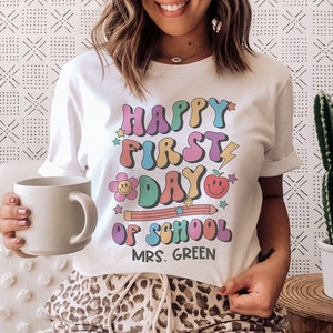 Puede incluir: Camiseta blanca con un gráfico colorido que dice "Happy First Day of School" con un arcoíris, una flor, un lápiz y una manzana. El texto "Mrs. Green" está debajo del gráfico.