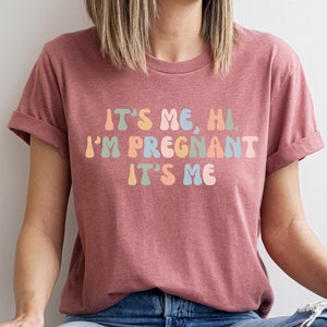 Puede incluir: Una mujer con una camiseta color marr&oacute;n rojizo con el texto "It's me, hi. I'm pregnant. It's me" en colores arco&iacute;ris.
