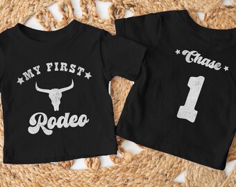 Pode incluir: Duas camisetas pretas com texto branco. Uma camiseta diz "My First Rodeo" com um gráfico de crânio de touro. A outra camiseta diz "Chase" com o número "1" abaixo.