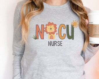 Può includere: Una maglietta grigia a maniche lunghe con il testo "NICU NURSE" in lettere colorate. Un leone di cartone animato con un fiore è al centro del testo. Un sole è a destra del testo.