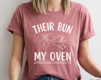 May include: A woman wearing a maroon t-shirt with the text "Their Bun My Oven #ProudSurrogate" in white. She is sitting with her legs crossed and wearing blue jeans.