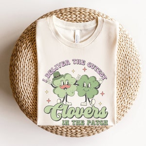 Puede incluir: Una camiseta beige con un diseño de trébol verde y el texto "I deliver the cutest clovers in the patch".