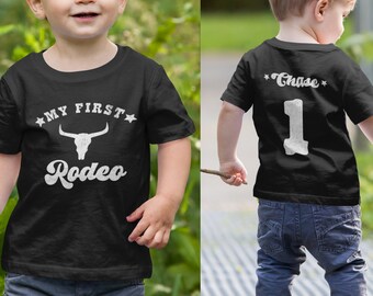 Pode incluir: Camiseta preta com texto branco que diz "My First Rodeo" e um gráfico de crânio de touro. Outra camiseta preta com texto branco que diz "Chase" e o número "1".