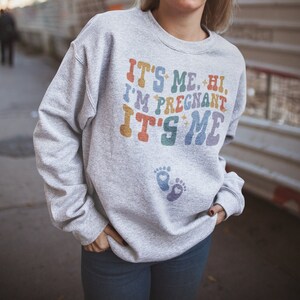 May include: A light gray sweatshirt with the text "IT'S ME, HI, I'M PREGNANT IT'S ME" in colorful retro lettering. Below the text are two baby footprints. The sweatshirt is a casual style.