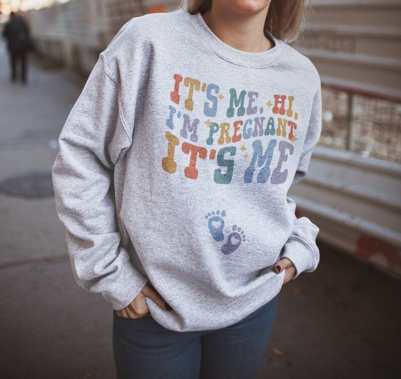 May include: A light gray sweatshirt with the text "IT'S ME, HI, I'M PREGNANT IT'S ME" in colorful retro lettering. Below the text are two baby footprints. The sweatshirt is a casual style.