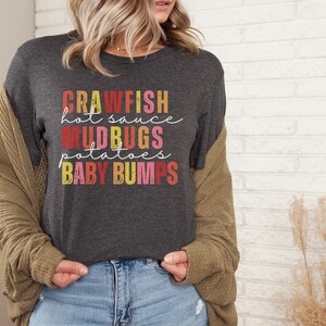Puede incluir: Una camiseta gris oscuro con un gr&aacute;fico de estilo vintage que dice "Crawfish hot sauce Mudbugs potatoes Baby Bumps".
