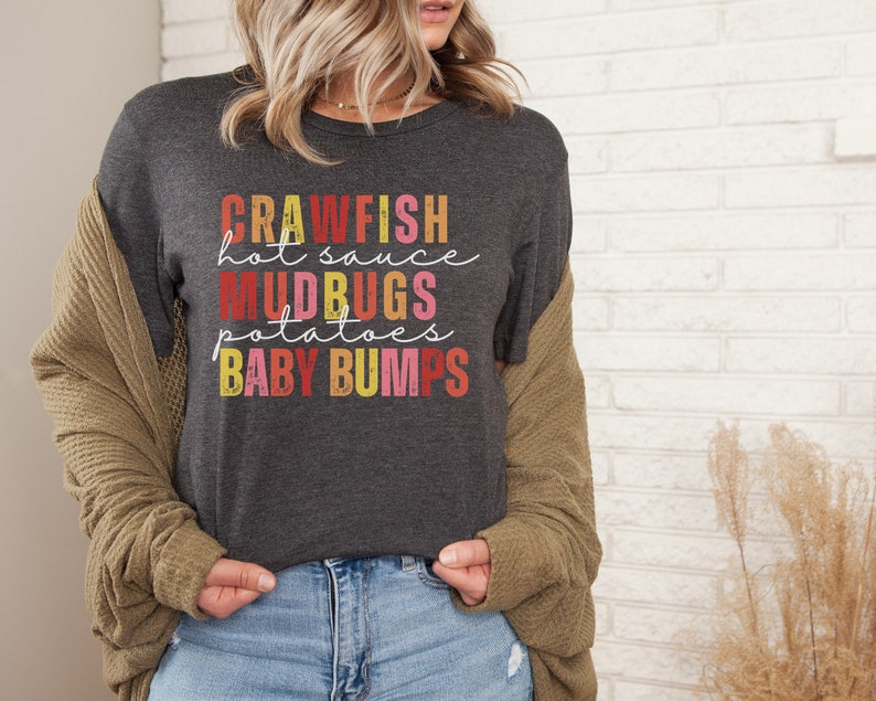 Puede incluir: Una camiseta gris oscuro con un gr&aacute;fico de estilo vintage que dice "Crawfish hot sauce Mudbugs potatoes Baby Bumps".