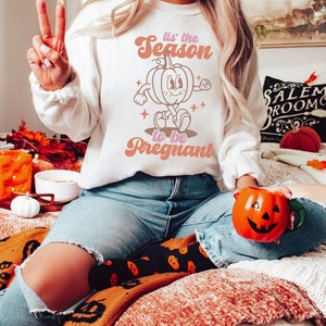 May include: A white sweatshirt with a retro-style graphic of a pumpkin with the text "Tis' the Season to be Pregnant".