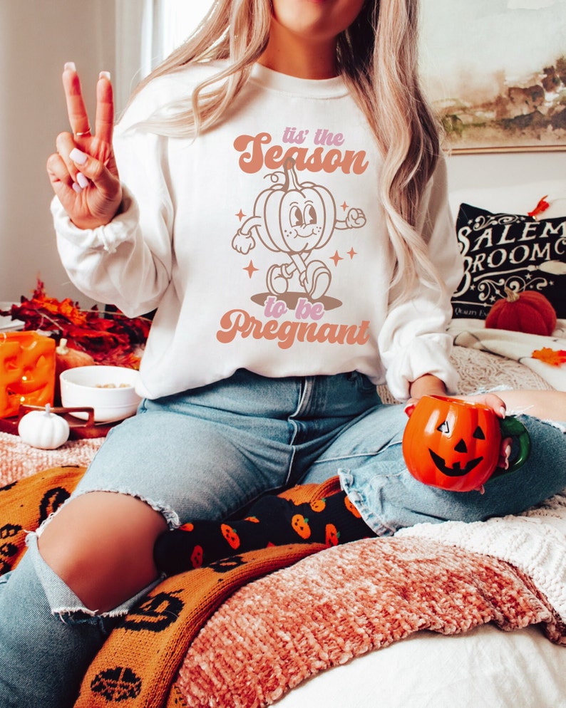 May include: A white sweatshirt with a retro-style graphic of a pumpkin with the text "Tis' the Season to be Pregnant".