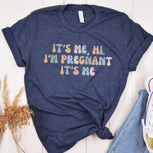 Puede incluir: Una camiseta azul marino con el texto "It's me, hi. I'm pregnant. It's me" en colores arco&iacute;ris.
