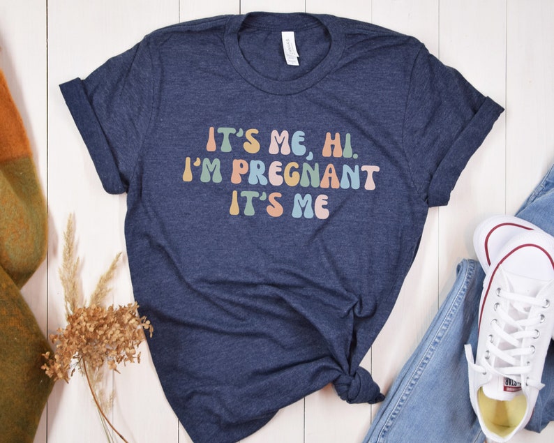 Puede incluir: Una camiseta azul marino con el texto "It's me, hi. I'm pregnant. It's me" en colores arco&iacute;ris.