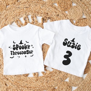 Op de afbeelding: Twee witte T-shirts met zwarte Halloween-themaontwerpen. E&eacute;n shirt zegt "Spooky Three-nager" met een heksenhoed en vleermuizen. Het andere shirt zegt "Suzie 3" met een heksenhoed.