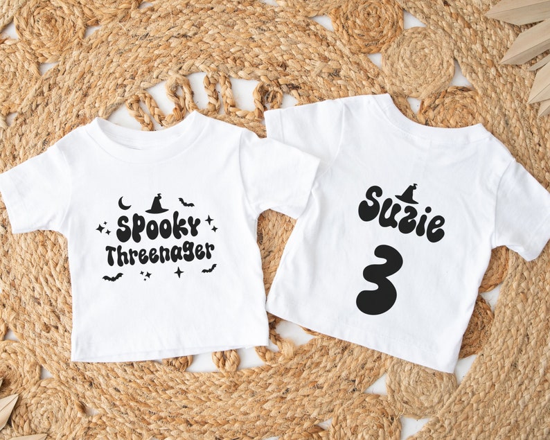 Op de afbeelding: Twee witte T-shirts met zwarte Halloween-themaontwerpen. E&eacute;n shirt zegt "Spooky Three-nager" met een heksenhoed en vleermuizen. Het andere shirt zegt "Suzie 3" met een heksenhoed.