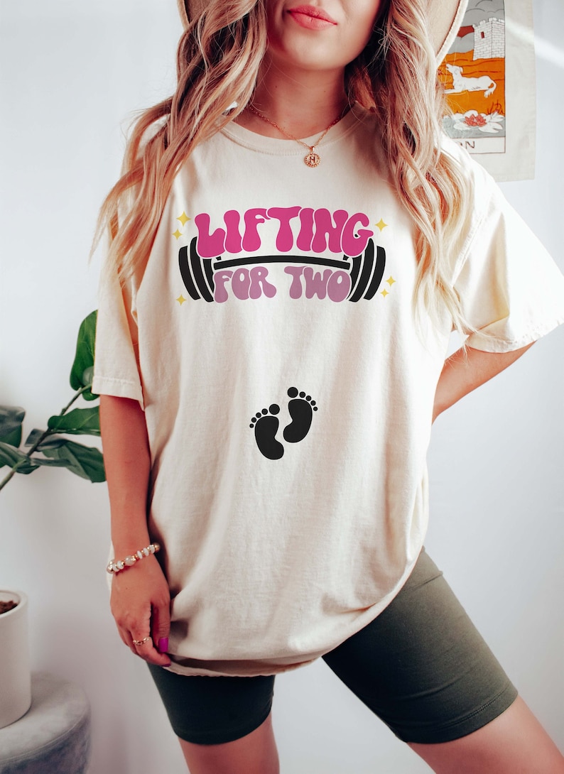 May include: A white t-shirt with the text "Lifting for Two" in pink and black lettering with a barbell graphic.  Two black baby footprints are below the text.