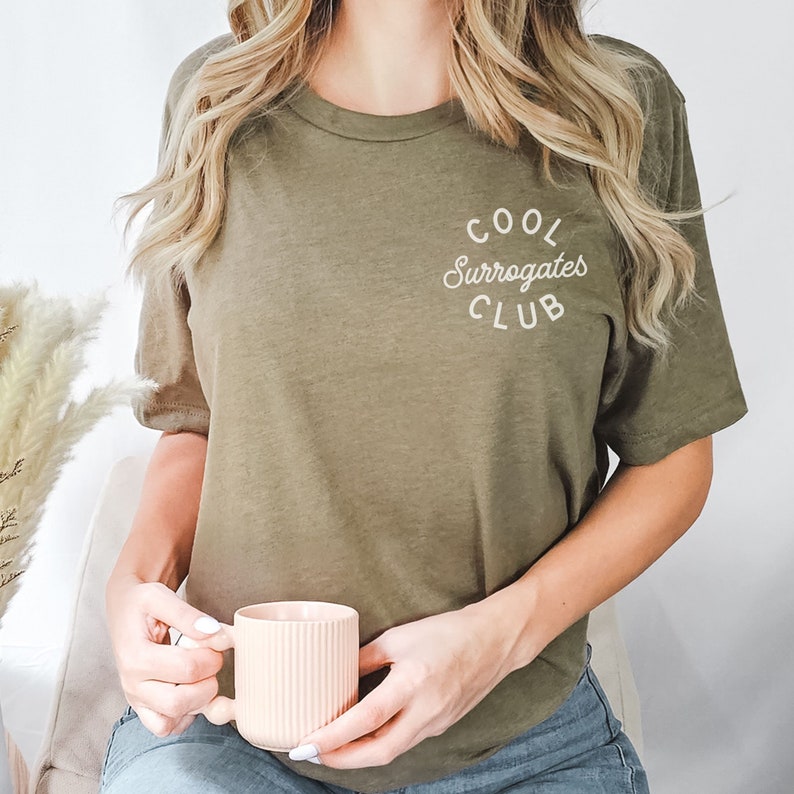 May include: Olive green t-shirt with the text "COOL Surrogates CLUB" in white script. The shirt is being held by a person wearing blue jeans and holding a pink mug with vertical ridges.
