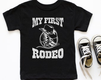 Pode incluir: Camiseta preta com um gráfico branco de uma bota de cowboy e um chapéu com o texto "MY FIRST RODEO".