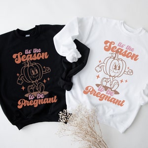 May include: Two sweatshirts featuring a pumpkin graphic and the text "tis' the season to be pregnant". One sweatshirt is black and the other is white.