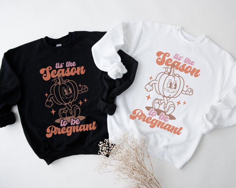 May include: Two sweatshirts featuring a pumpkin graphic and the text "tis' the season to be pregnant". One sweatshirt is black and the other is white.