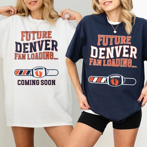 以下が含まれることがあります： 白とネイビーブルーの2枚のTシャツには、「FUTURE DENVER FAN LOADING... COMING SOON」の文字がプリントされています。デザインにはフットボールと赤ちゃんの足跡が含まれています。妊娠の発表用Tシャツです。