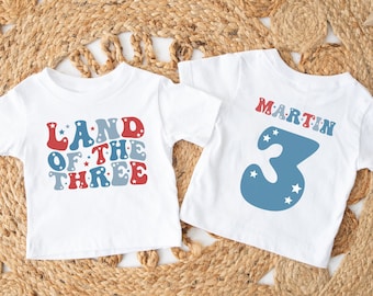 Könnte beinhalten: Zwei weiße T-Shirts mit rot-weiß-blauem Text und einer blauen Zahl 3. Das erste Shirt sagt "Land of the Three" und das zweite Shirt sagt "Martin" über der Zahl 3.