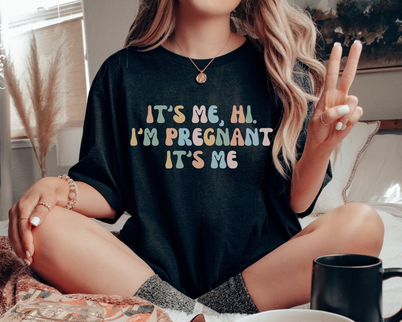 Puede incluir: Una camiseta negra con el texto "It's me, hi. I'm pregnant. It's me" en colores arco&iacute;ris.