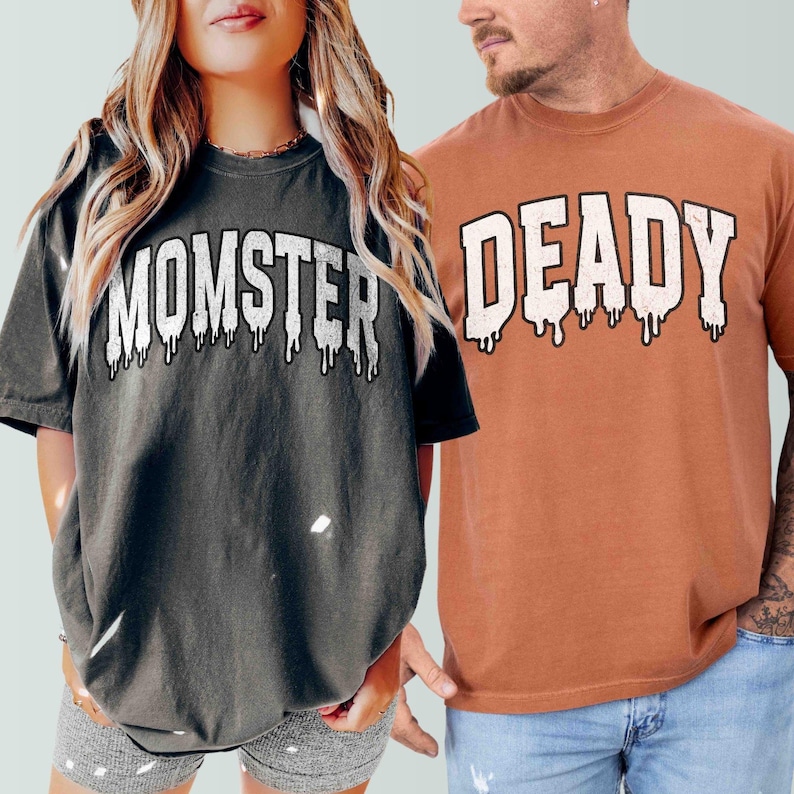 May include: Two t-shirts with a Halloween theme. The first shirt is dark gray with the text "MOMSTER" in white dripping letters. The second shirt is orange with the text "DEADLY" in white dripping letters.