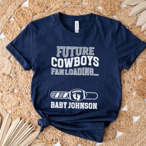 Puede incluir: Una camiseta azul marino con el texto "Future Cowboys Fan Loading..." y una barra de progreso con un gr&aacute;fico de f&uacute;tbol. El texto "Baby Johnson" est&aacute; debajo de la barra de progreso.