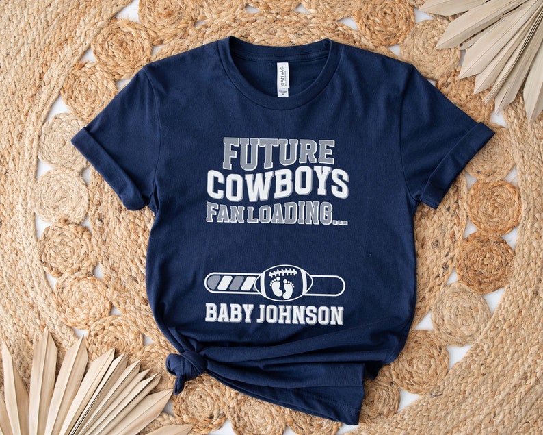 Puede incluir: Una camiseta azul marino con el texto "Future Cowboys Fan Loading..." y una barra de progreso con un gr&aacute;fico de f&uacute;tbol. El texto "Baby Johnson" est&aacute; debajo de la barra de progreso.