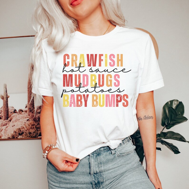 Puede incluir: Camiseta blanca con un dise&ntilde;o gr&aacute;fico colorido que dice "Crawfish hot sauce Mudbugs potatoes Baby Bumps".