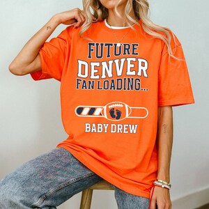 以下が含まれることがあります： オレンジ色のTシャツ。白とネイビーブルーで「FUTURE DENVER FAN LOADING...」の文字。その下に、赤ちゃんの足跡が付いたフットボールと「BABY DREW」の名前。妊娠発表用のTシャツです。