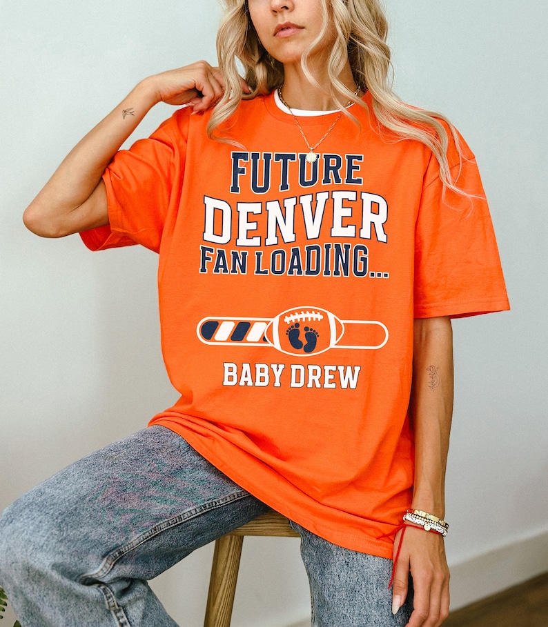 以下が含まれることがあります： オレンジ色のTシャツ。白とネイビーブルーで「FUTURE DENVER FAN LOADING...」の文字。その下に、赤ちゃんの足跡が付いたフットボールと「BABY DREW」の名前。妊娠発表用のTシャツです。