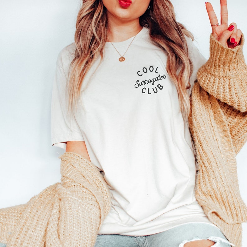May include: Cream-colored t-shirt with the text "COOL Surrogates CLUB" in a curved, black font. The shirt is paired with a tan sweater draped over the shoulders. A gold necklace is visible.