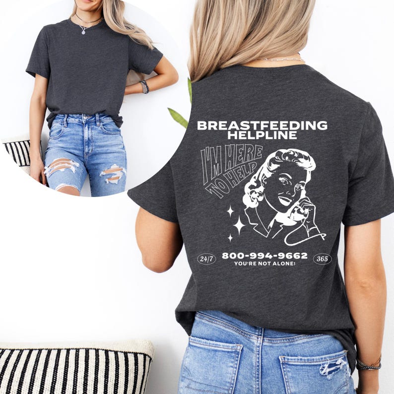 May include: A dark grey t-shirt with a white graphic of a woman on the phone. The graphic says "I'm here to help" and includes the text "Breastfeeding Helpline 800-994-9662 You're not alone!" with the numbers 24/7 and 365.