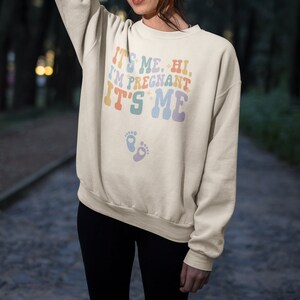 May include: Cream-colored sweatshirt with the text "IT'S ME. HI. I'M PREGNANT IT'S ME" in pastel colors. Below the text are two baby footprints. The sweatshirt is a relaxed fit.