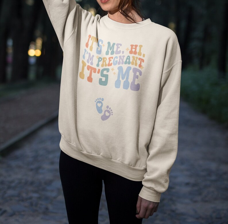 May include: Cream-colored sweatshirt with the text "IT'S ME. HI. I'M PREGNANT IT'S ME" in pastel colors. Below the text are two baby footprints. The sweatshirt is a relaxed fit.