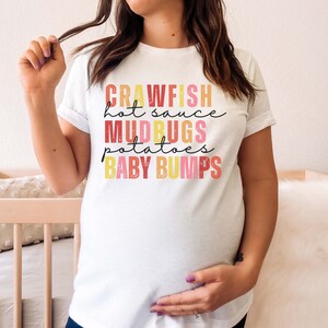 Puede incluir: Camiseta blanca con un gr&aacute;fico colorido que dice "Crawfish hot sauce Mudbugs potatoes Baby bumps".