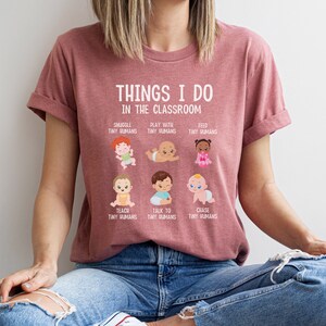 May include: A pink t-shirt with the text "Things I do in the classroom" and illustrations of babies. The text below the illustrations includes phrases like "Snuggle tiny humans", "Play with tiny humans", "Feed tiny humans", "Teach tiny humans", "Talk to tiny humans", and "Chase tiny humans".