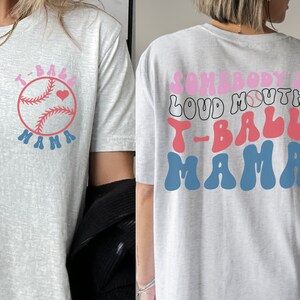 May include: Gray t-shirt with a pink and blue baseball graphic and the text "T-Ball Mama" on the front. The back of the shirt has the text "Somebody's Loud Mouth T-Ball Mama" in pink, red, and blue.