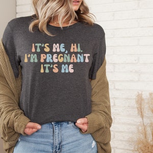 Puede incluir: Una camiseta gris oscuro con el texto "It's me, hi. I'm pregnant. It's me" en colores arco&iacute;ris.