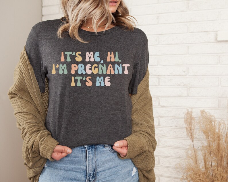 Puede incluir: Una camiseta gris oscuro con el texto "It's me, hi. I'm pregnant. It's me" en colores arco&iacute;ris.