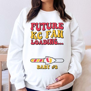 May include: White sweatshirt with the text "FUTURE KC FAN LOADING..." in red and yellow letters. A progress bar with a red and yellow arrow and the text "BABY #2" is below the text.