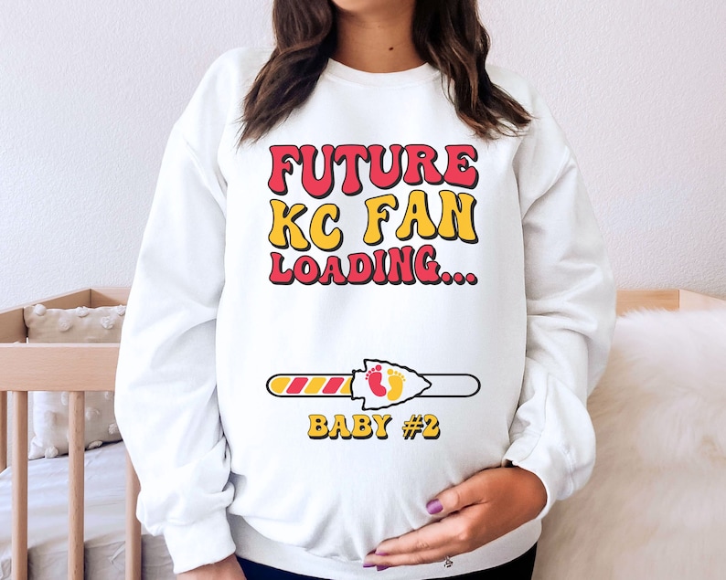 May include: White sweatshirt with the text "FUTURE KC FAN LOADING..." in red and yellow letters. A progress bar with a red and yellow arrow and the text "BABY #2" is below the text.