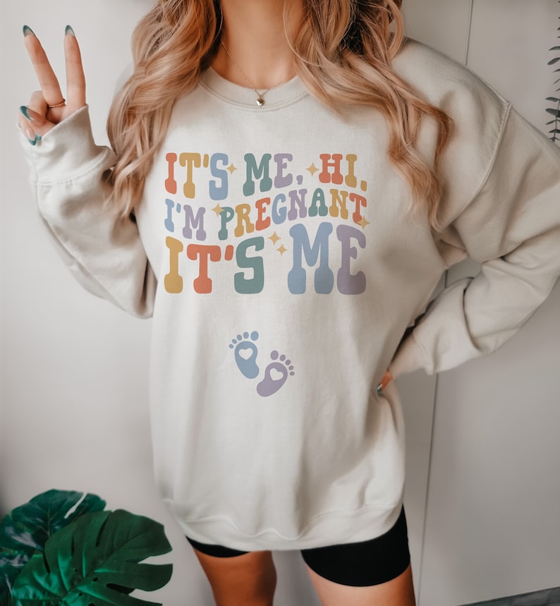 May include: Beige sweatshirt with the text "IT'S ME, HI, I'M PREGNANT IT'S ME" in colorful retro font. Below the text are two baby footprints with hearts. The sweatshirt has long sleeves.