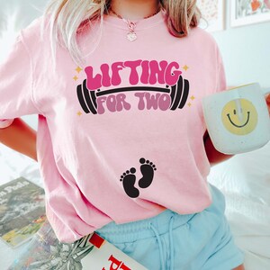 May include: Pink t-shirt with the text "Lifting for Two" in a retro font with a barbell and two baby footprints.