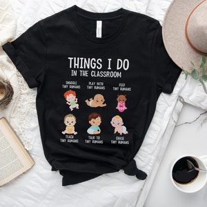 May include: Black t-shirt with a white graphic that says "Things I do in the classroom" and features illustrations of babies with text describing different activities like "Snuggle tiny humans", "Play with tiny humans", "Feed tiny humans", "Teach tiny humans", "Talk to tiny humans", and "Chase tiny humans".
