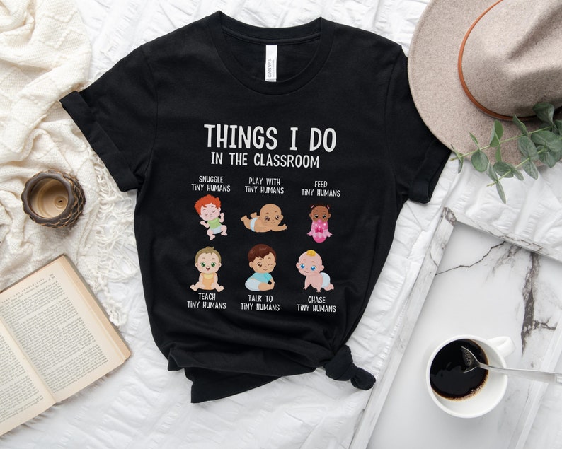 May include: Black t-shirt with a white graphic that says "Things I do in the classroom" and features illustrations of babies with text describing different activities like "Snuggle tiny humans", "Play with tiny humans", "Feed tiny humans", "Teach tiny humans", "Talk to tiny humans", and "Chase tiny humans".